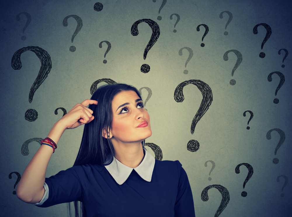 Portrait confused thinking young woman bewildered scratching head seeks a solution looking up at many question marks isolated on gray wall background. Human face expression