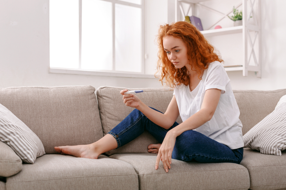 Worried redhead girl checking her recent pregnancy test, sitting on beige couch at home