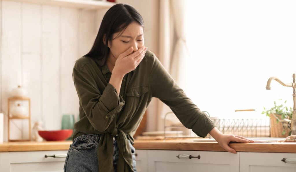 Young asian woman in casual feeling nausea, cover mouth, standing next to table in kitchen, copy space. Toxicosis during pregnancy, food poisoning concep
