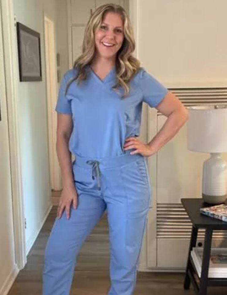 A nurse standing in her scrubs. A table, lamp, walls, and wooden floor in the background. 