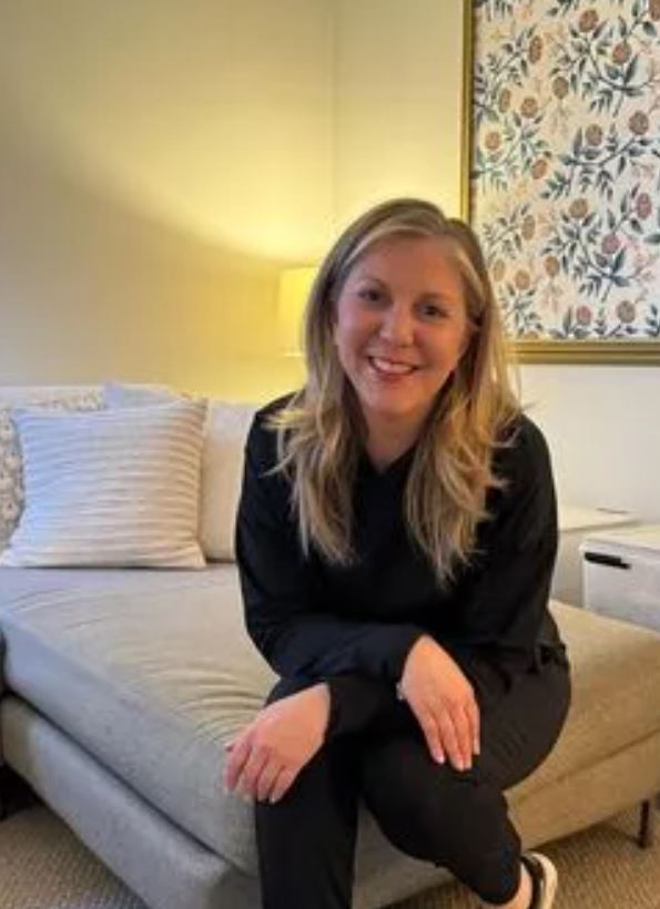 A woman wearing black, sitting at the end of a couch. A floral canvas, lamp, and light-colored furniture in the background. 