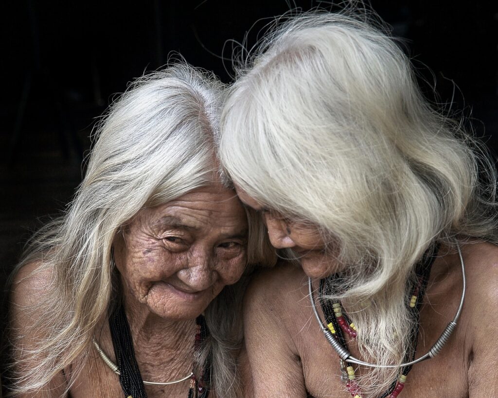 Two older women with white and gray hair sitting side by side. Black background,