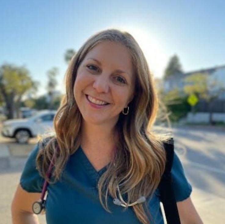 A nurse in her scrubs with a stethoscope. Blurred background with blue skies, trees, and vehicles. 