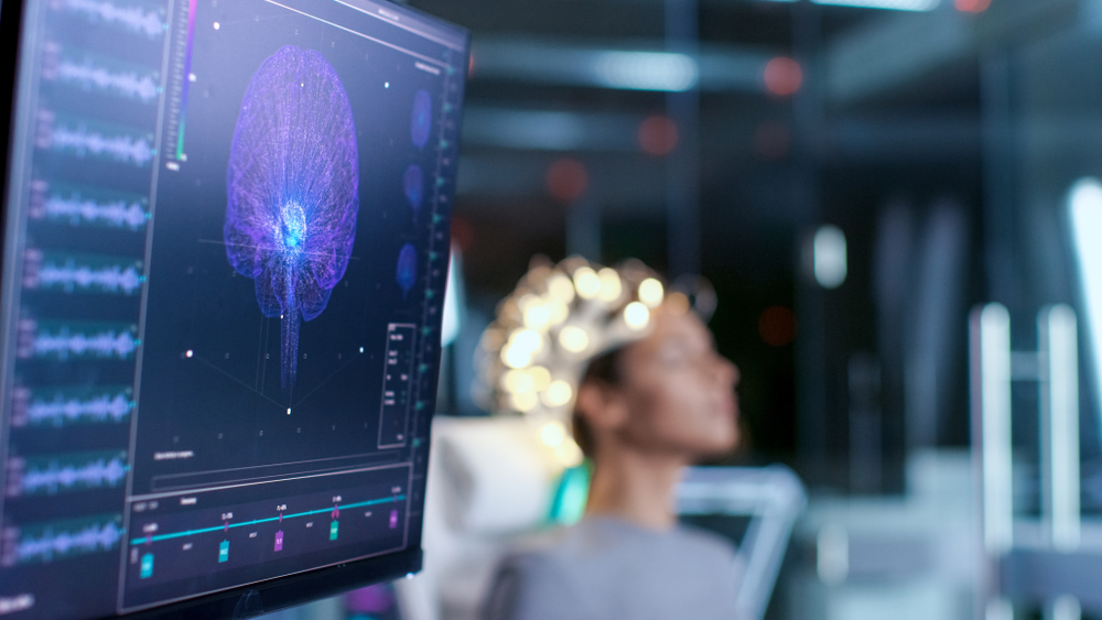 Woman Wearing Brainwave Scanning Headset Sits in a Chair In the Modern Brain Study Laboratory/ Neurological Research Center. Monitors Show EEG Reading and Brain Model.
