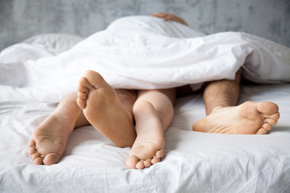 Close up of passionate lovers feet touch having sex on white sheet in bedroom, sensual couple enjoy intimate moment relaxing in bed, legs of boyfriend and girlfriend make love in woman on top position
