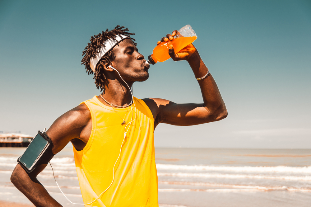 Black runner taking rest drinking energy drink at the beach at summer
