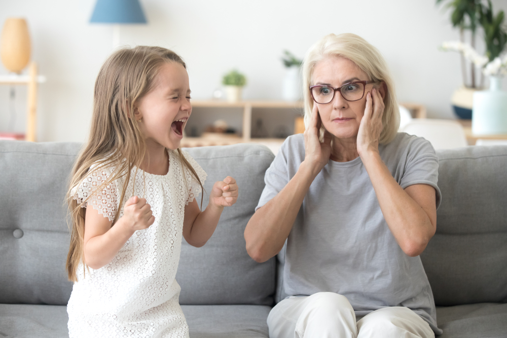 Shocked grandma closing ears not to hear noisy stubborn fussy little granddaughter screaming demanding attention, preschool spoiled kid girl yelling at grandmother, child tantrum manipulation concept
