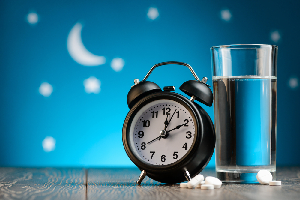 Alarm clock, pills and glass of water on a bedside table. Insomnia, treatment for sleepless nights.