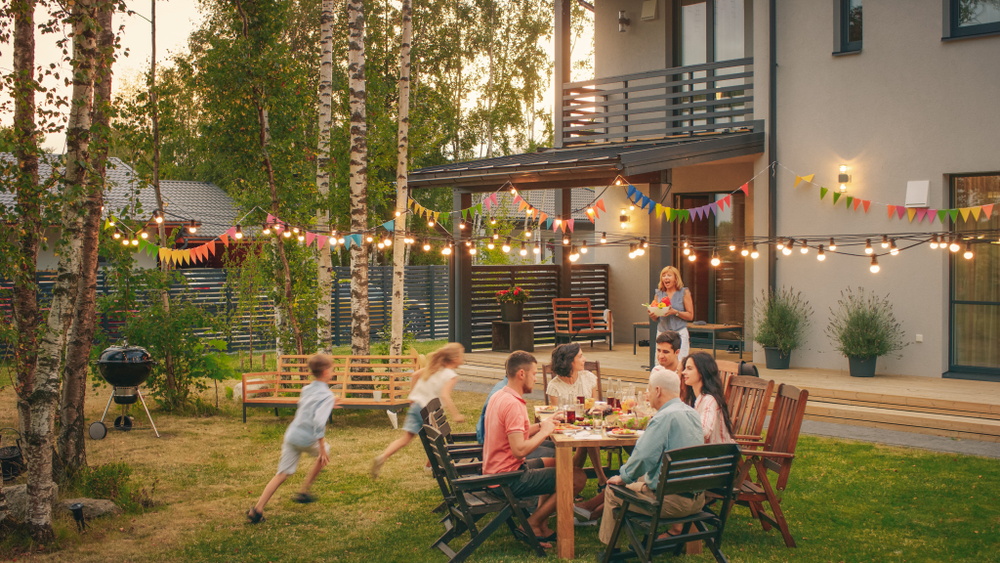 Big Family Garden Party Celebration, Gathered Together at the Table Family, Friends and Children. People are Drinking, Passing Dishes, Joking and Having Fun. Kids Run Around Table.
