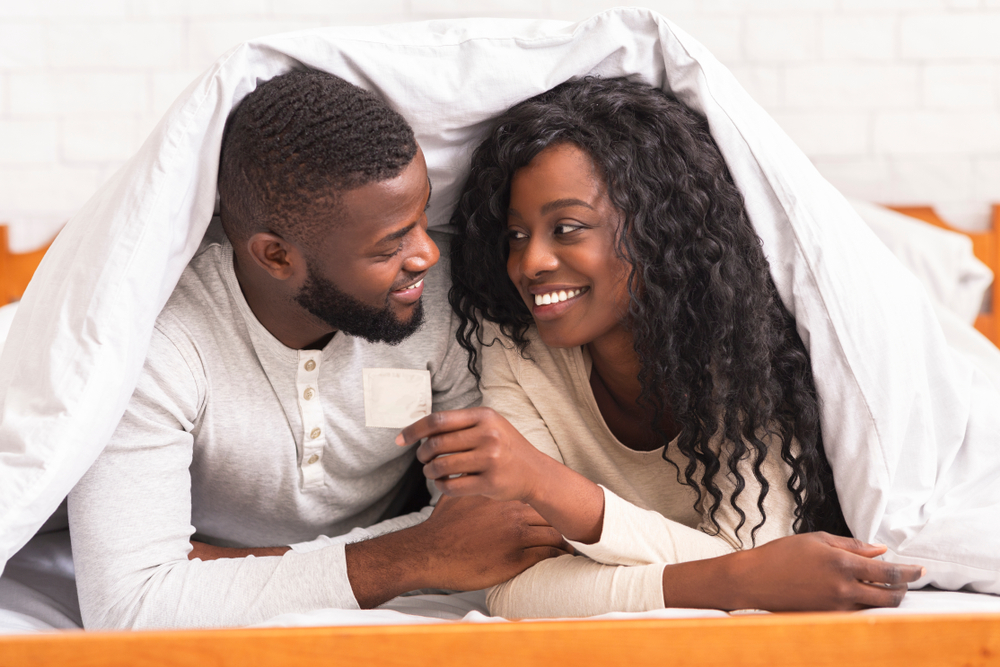 Safe sex. Young african american couple holding condom, lying on bed under white blanket, looking at each other with love and care
