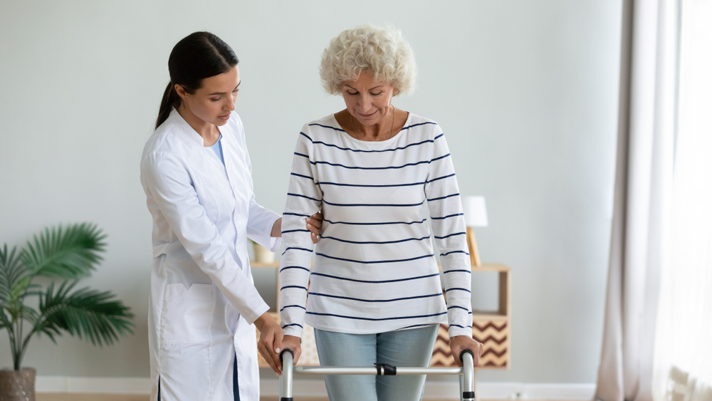 Caregiver teaching disabled older woman to use walker at home, training, recovery and rehabilitation after injury, girl nurse wearing white uniform holding mature patient hand, supporting
