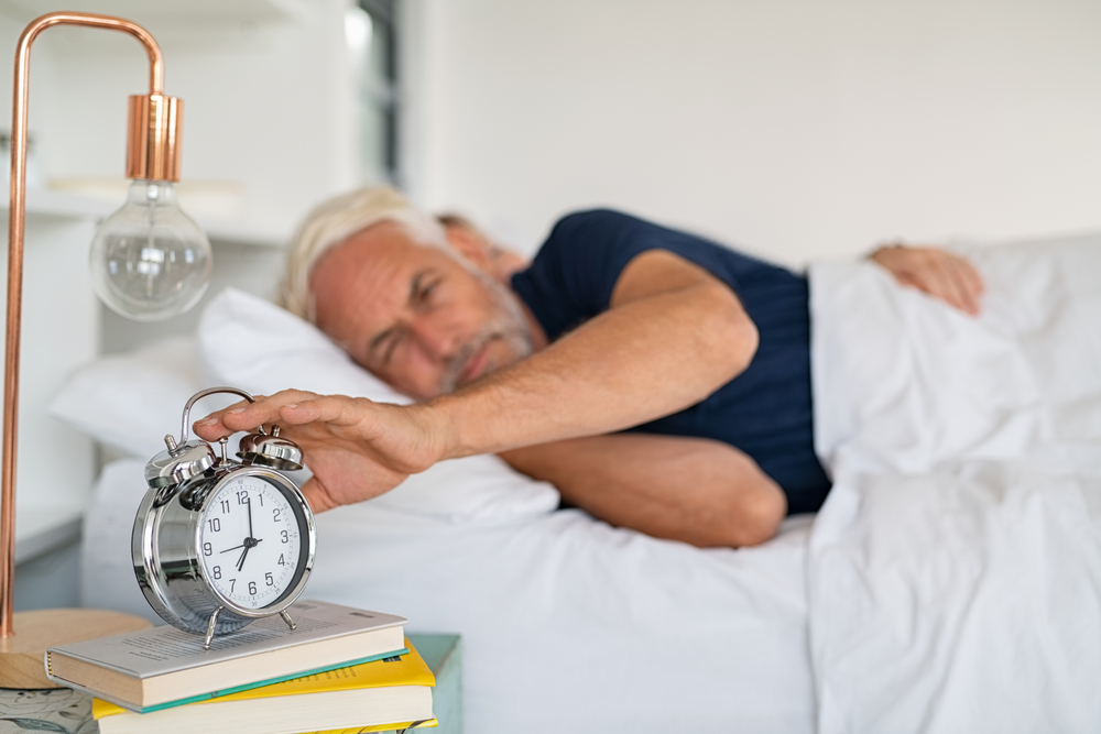 Old sleepy man lying on bed and stopping alarm clock. Senior man lying on side on bed turning off an alarm clock in the morning at 7am. Mature adult waking up in the morning.
