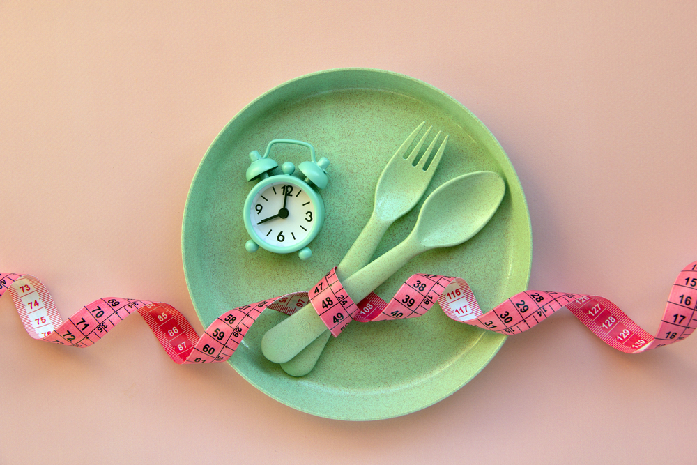 Creative flat lay composition with plate, alarm clock, spoon, fork and measuring tape on pink background. Intermittent fasting, ketogenic, diet concept.  Flat lay, copy space.

