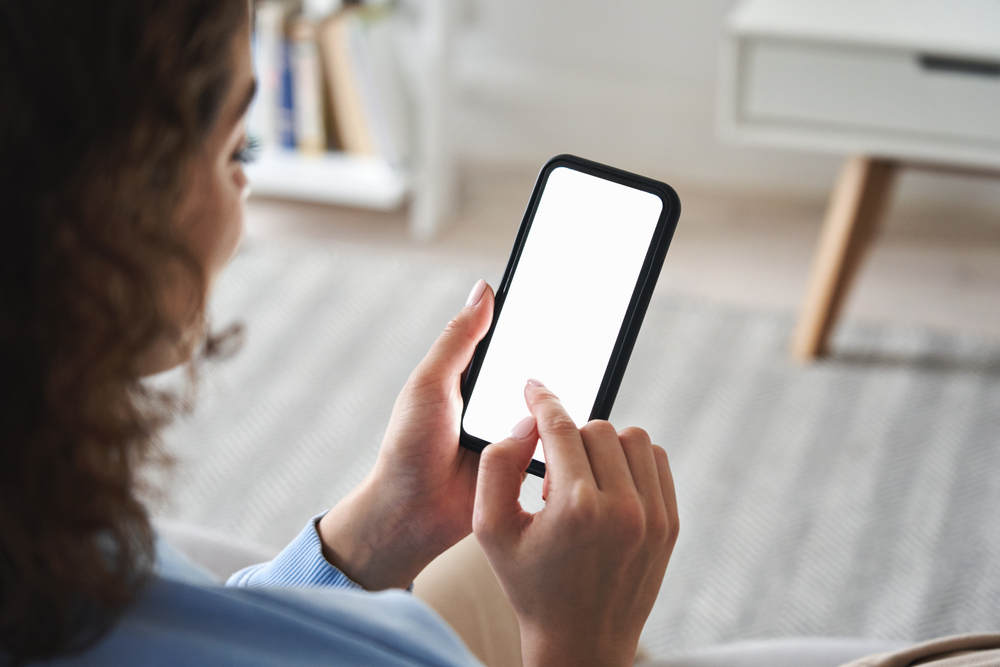 Young woman or teen girl hands holding cell phone touching finger mockup white blank display, empty screen for social media app ad at home. Mobile application tech concept, over shoulder closeup view.
