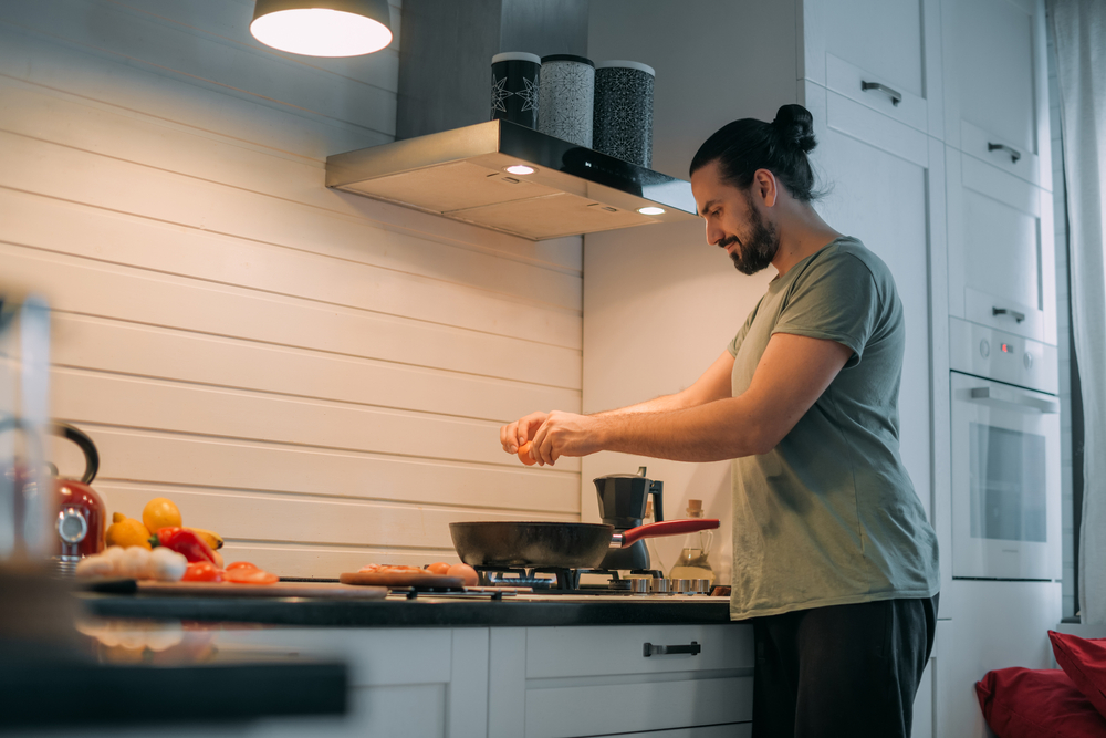 A man prepares breakfast in the kitchen. Young handsome caucasian male preparing food for himself for lunch on a gas stove in a large bright kitchen