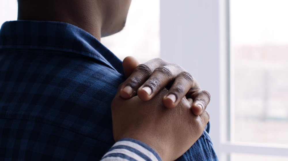 Sad black man looking at window at home during pandemic outbreak, his wife touching his shoulder, comforting him, back view, slow motion
