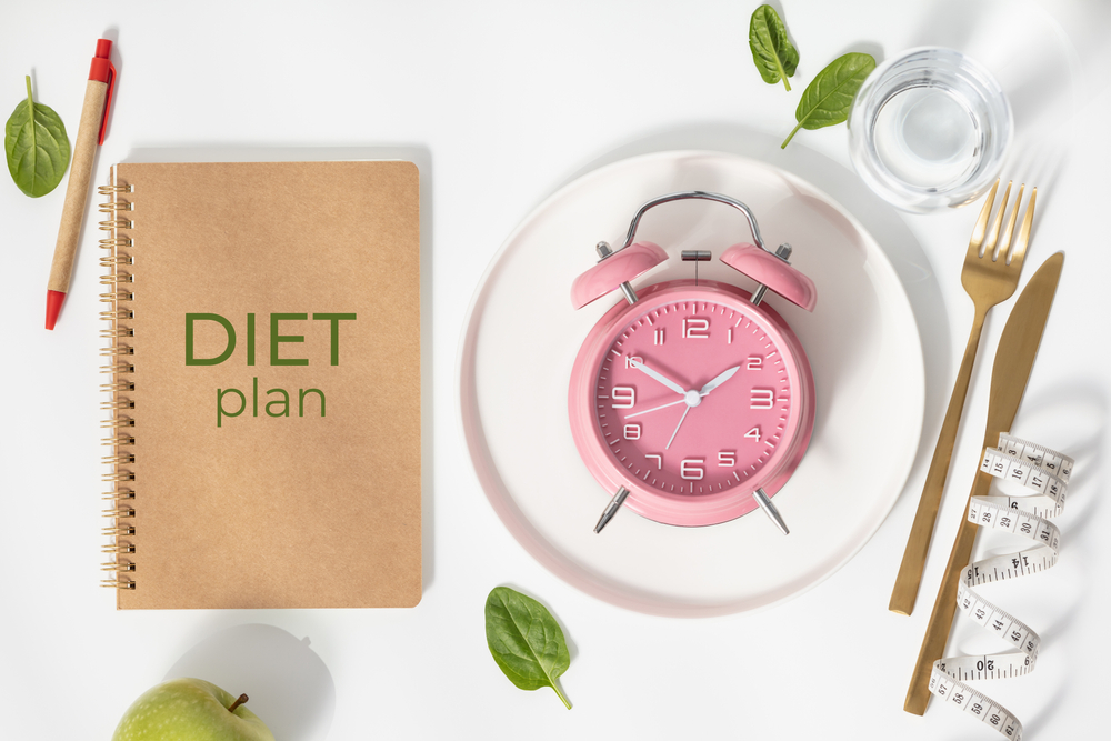 Composition with cutlery, plate, measuring tape, paper notebook and alarm clock on white background, flat lay, top view, copy space