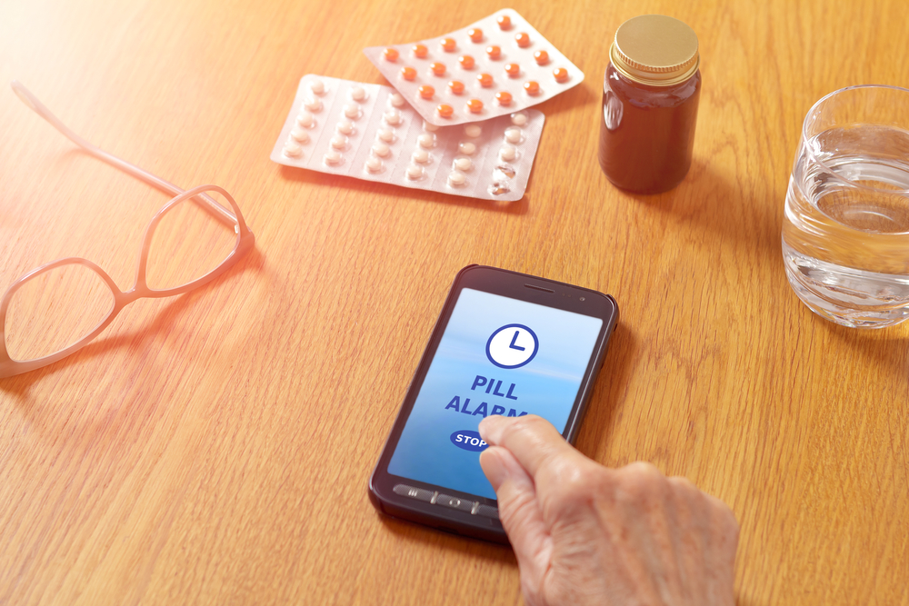 Pill alarm app on mobile, cell or smartphone, with water glass, eyeglasses, medicine and the hand of a senior woman.