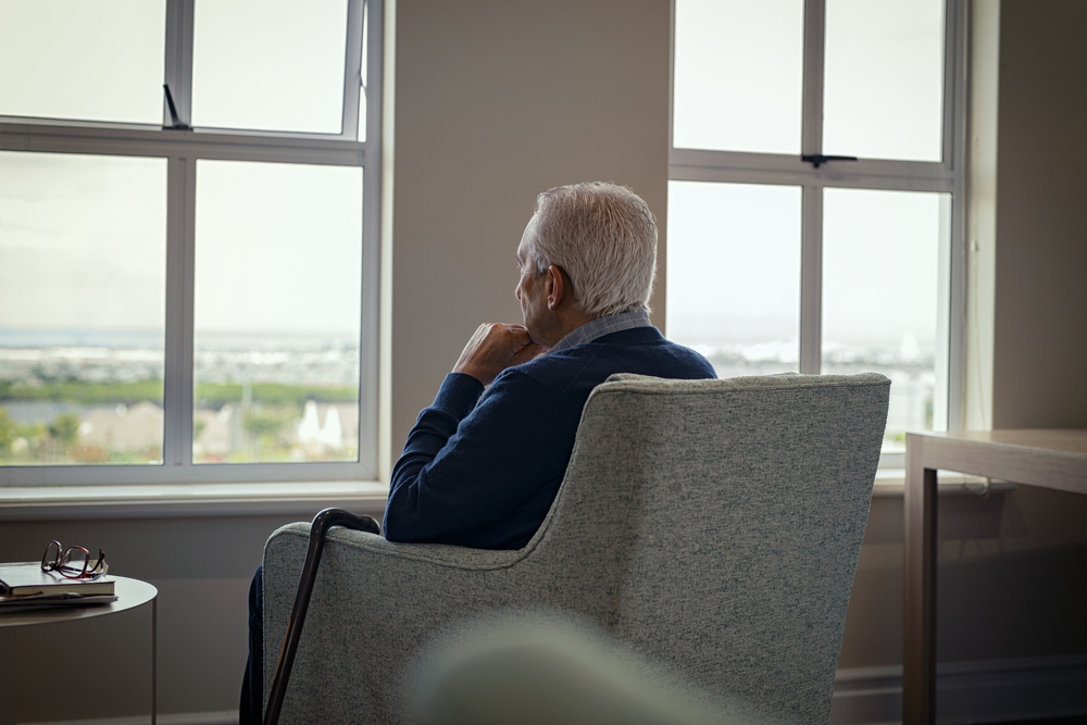 Rear view of senior man sitting on armchair and looking through the window. Lonely old man sitting at home near window during covid19 outbreak. Thoughtful retired man abandoned at nursing home.
