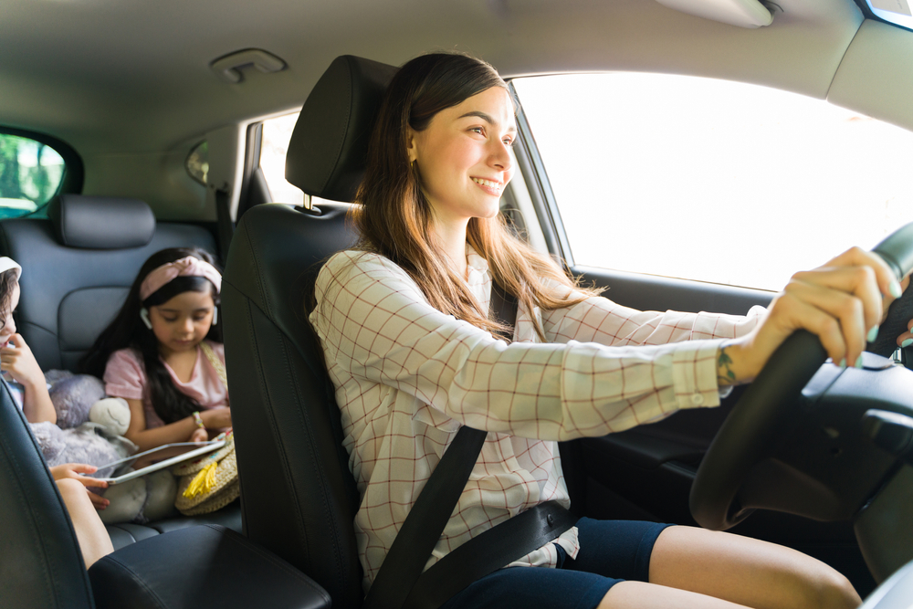 Entertaining my children. Beautiful young mom smiling while driving a car with her kids in the back watching a movie on a tablet with headphones during her errands