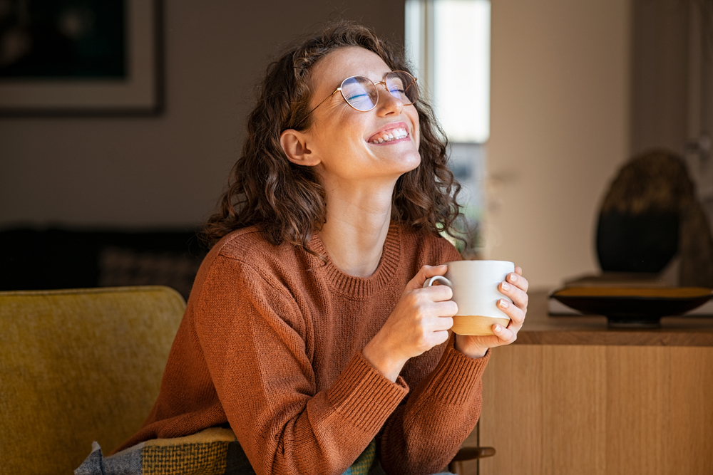 Portrait of joyful young woman enjoying a cup of coffee at home. Smiling pretty girl drinking hot tea in winter. Excited woman wearing spectacles and sweater and laughing in an autumn day.