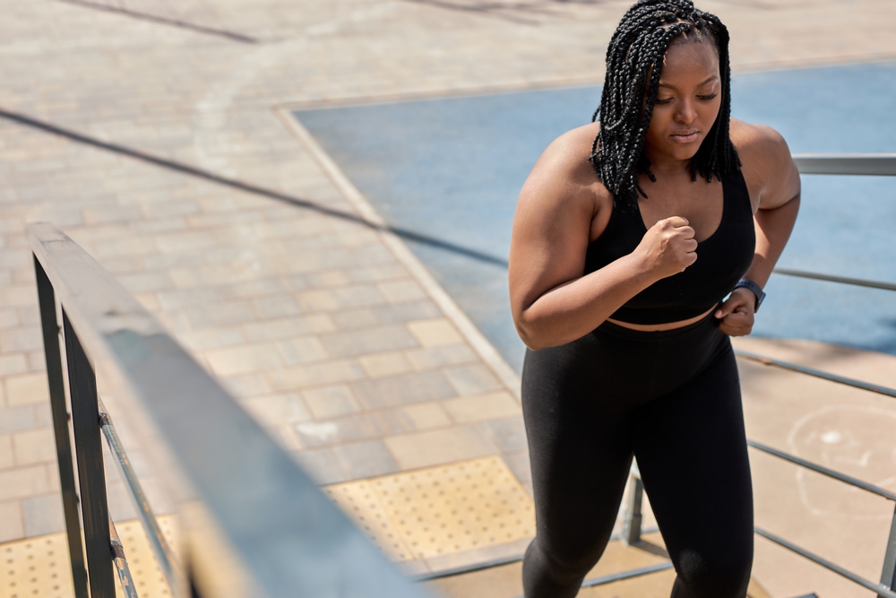 Obese young african woman foing fitness exercises on stairs outdoors. Weight loss cardio goal achievement challenge. Copy space. Side view portrait of black lady in black tracksuit engaged in fitness