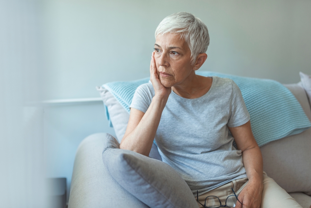 Upset depressed senior gray-haired lady touching temple, sitting on sofa, looking in the window, feeling worried and stressed about the future and thinking about own problems