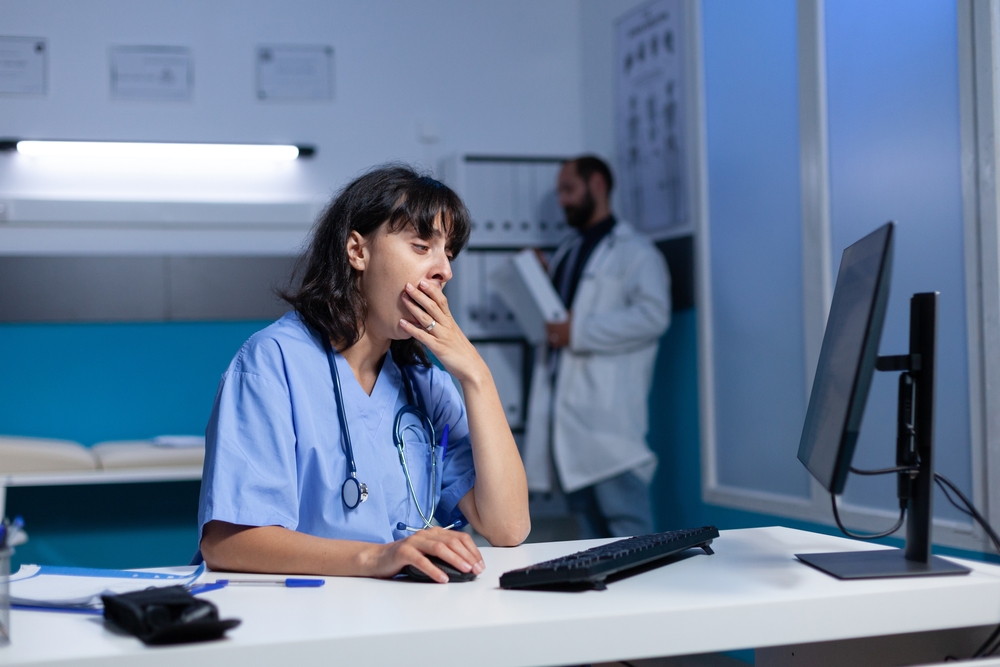 Exhausted nurse using computer on desk while falling asleep, working late. Medical assistant looking at monitor and feeling burnout after overtime work at night in healthcare office.
