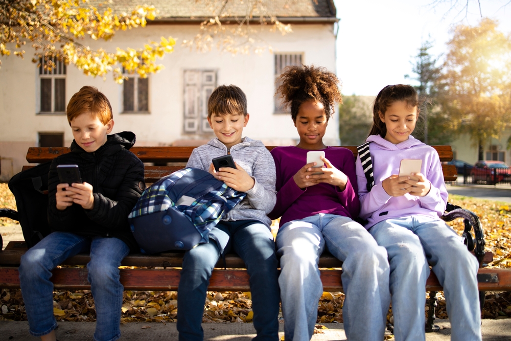 Group of multicultural children sitting on bench and using their smart phones surfing internet - technology concept.
