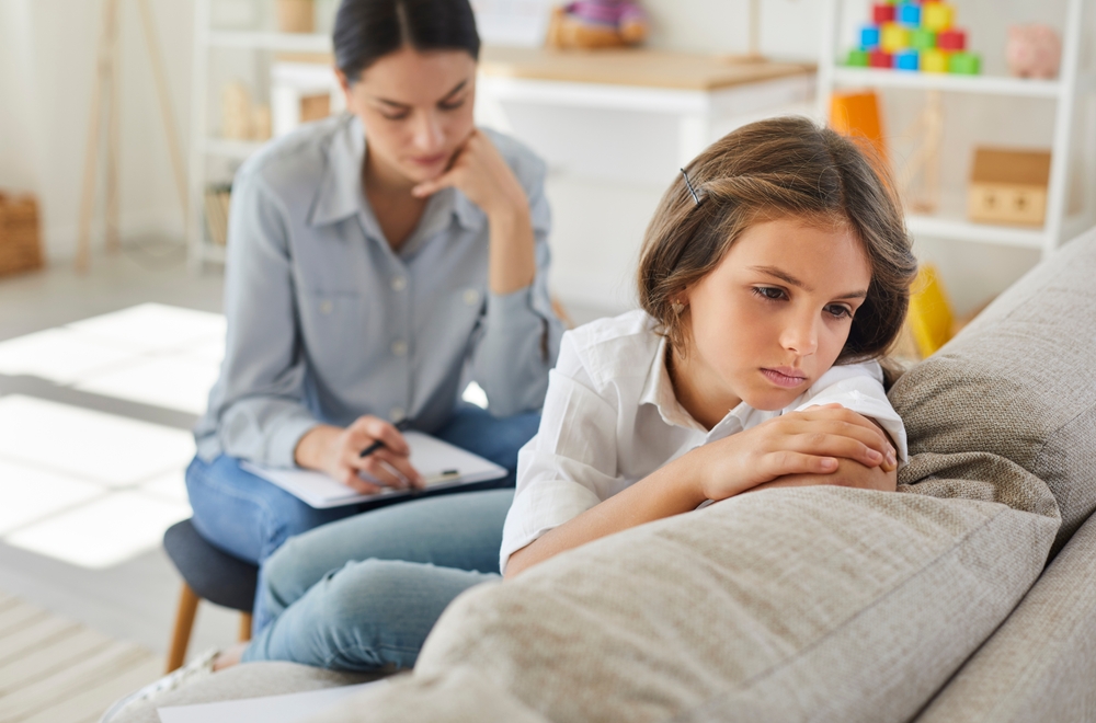 Frustrated little child at psychologist's office. Portrait of girl with sad face sitting on couch during therapy session. Therapist trying to help unhappy, resentful kid who has behaviour issues
