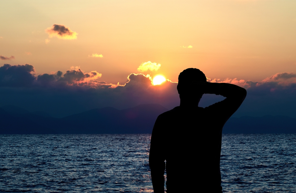 Back view silhouette photo of depressed man watching the sunseton the sea. Thoughtful, overthink, pensive and future plans concept.