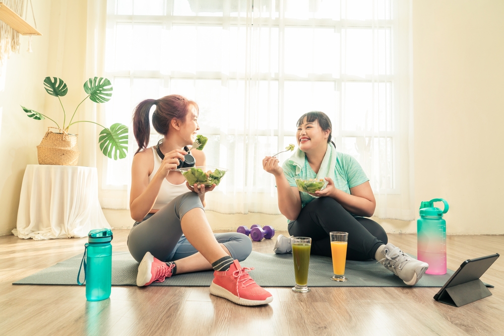 Two Asian women body size different in sport wear sitting healthy eating vegetable salad after exercise at home together. Dieting and exercise at home concept.