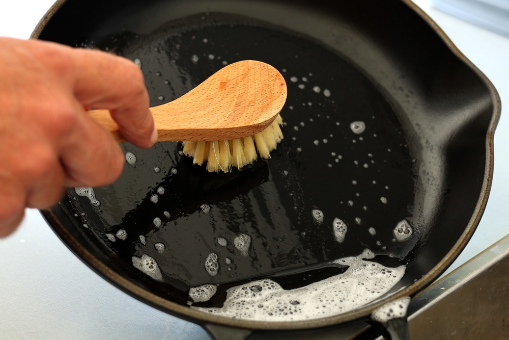 Close up A hand holding bamboo cleaning brush scrub and washing the dirty cast iron pan with a pattern of grease and oil stains.
