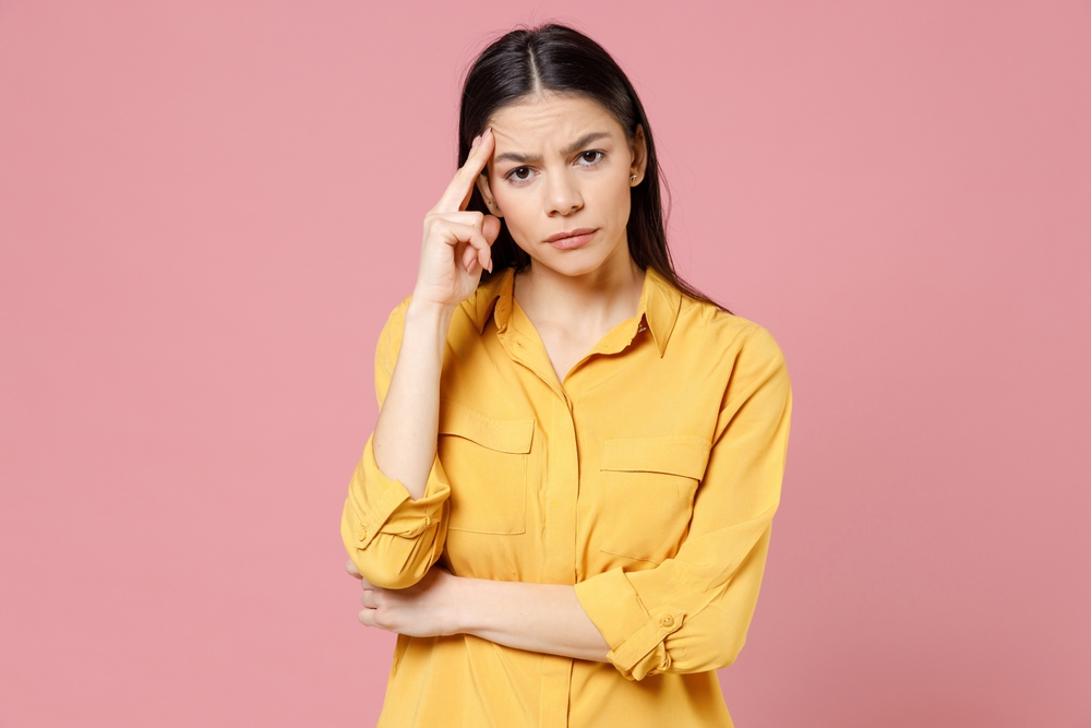 Young brunette thoughtful pensive overthinking latin student woman 20s wearing yellow casual shirt proping up forehead head looking camera isolated on pastel pink color background studio portrait