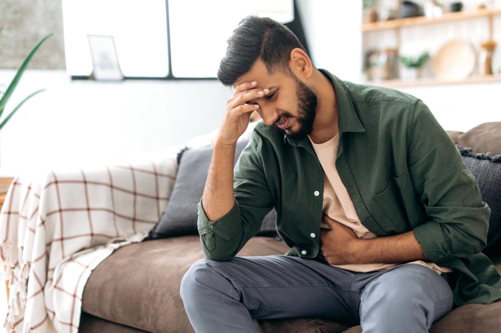 Unhappy indian or arabian man, sits on comfortable sofa in cozy living room, holds his hands on his stomach, grimaces from pain in his stomach, suffers from poisoning, spasm, stomach problems
