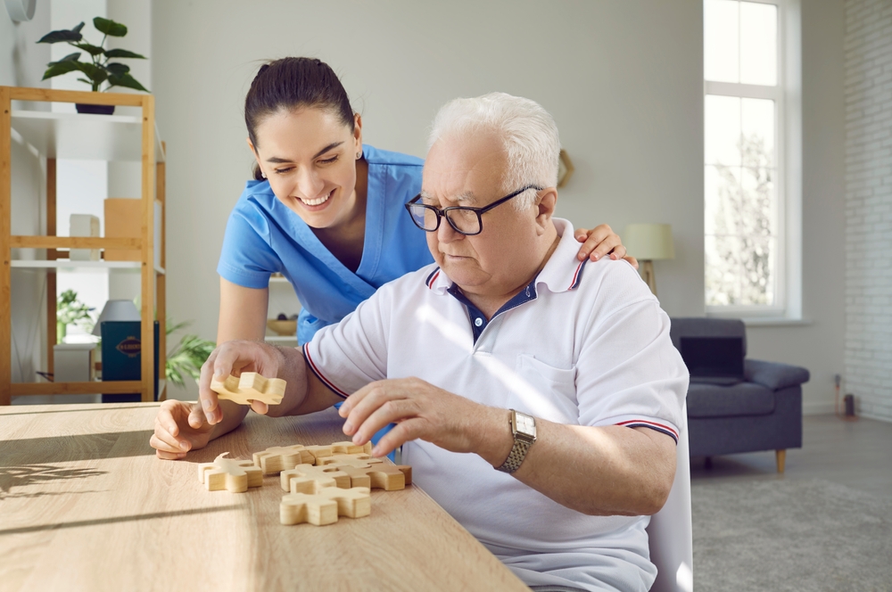 Nurse in retirement home helping old male patient with board game. Senior man with cognitive disorder sitting at table in geriatric clinic, playing games and thinking. Dementia, Alzheimers, care