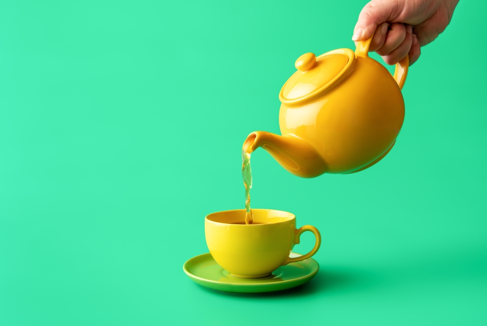 Woman's hand pour tea from yellow jug in a cup, minimalist on a green table. Hot healthy drink, mint tea in a yellow-colored cup.
