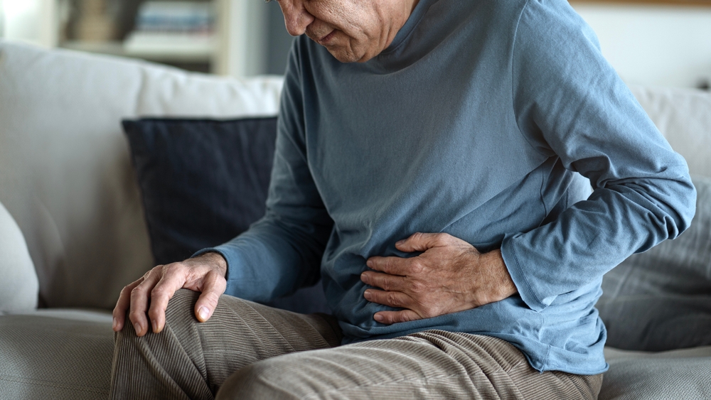 An elderly woman is sitting on the sofa at home, holding her stomach with her hands. Feels the pain of internal organs, indigestion, poisoning, menstrual pain.