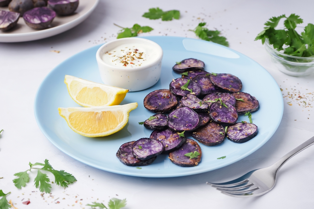 Plate with slices of fried purple potatoes, lemon and sauce on light background