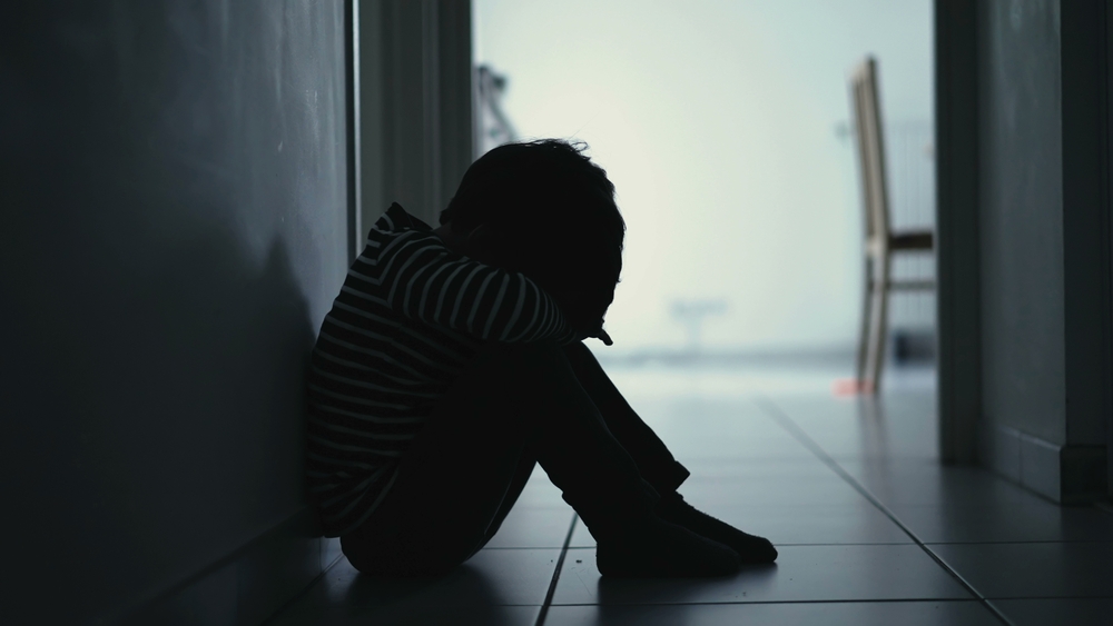 Sad child suffering from depression sitting alone in corridor feeling loneliness. Scared fearful small boy covering face in silhouette at home