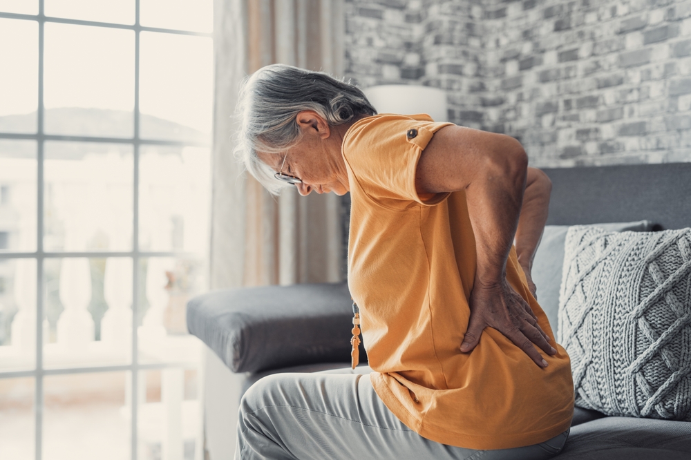 Unhappy mature woman touching back, feeling pain, sitting on couch in living room, unhealthy middle aged senior female suffering from backache, spinal problem, rubbing stiff muscles or kidney