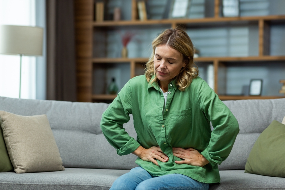 An elderly woman is sitting on the sofa at home, holding her stomach with her hands. Feels the pain of internal organs, indigestion, poisoning, menstrual pain.
