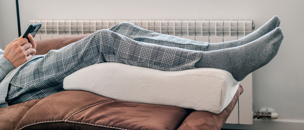 a man leans his legs on a leg elevation pillow made of memory foam, resting on the sofa, in a panoramic format to use as web banner or header