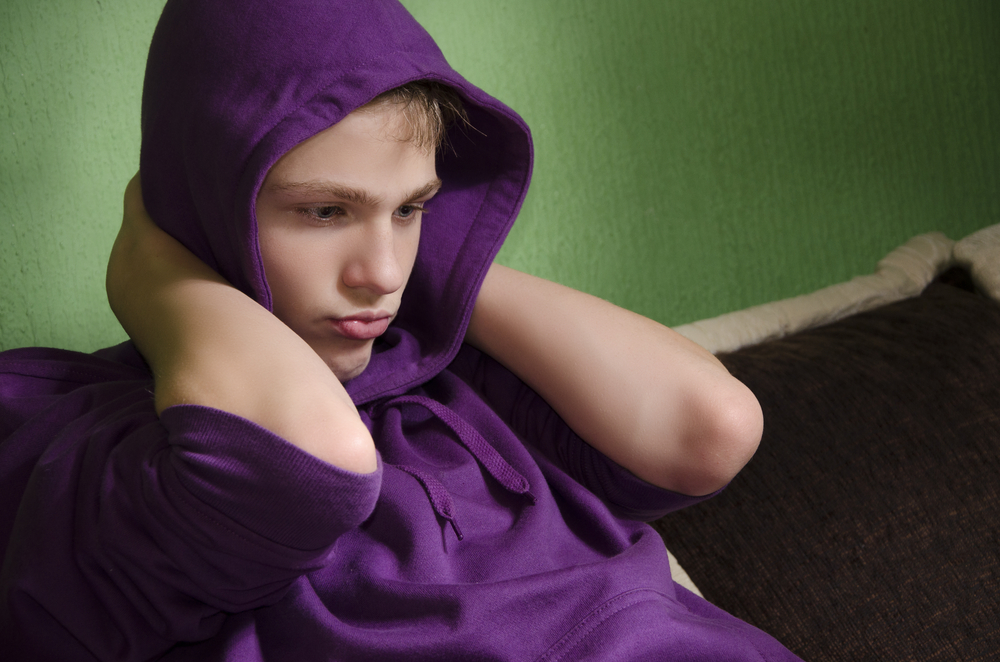 Depressed teenager with arms around his neck and hood on head, looking down.
