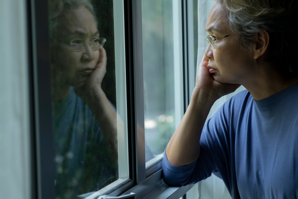 Portrait of sad and worried Asian senior woman looking through the window at home
