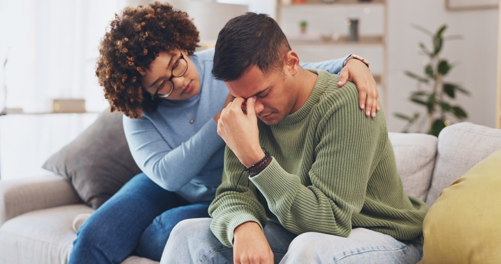 Grief, loss with mental health and sad, stress and people in relationship in crisis. Couple, depression and woman comfort man with empathy, support and love while sitting in living room at home.
