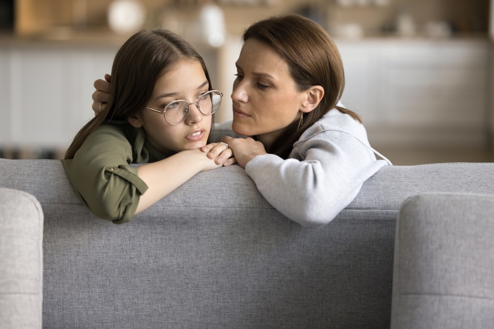 Serious empathetic mom listening to sad frustrated teenage daughter kid at home, giving comfort, support, compassion, love to child, discussing kids problems, bulling on sofa
