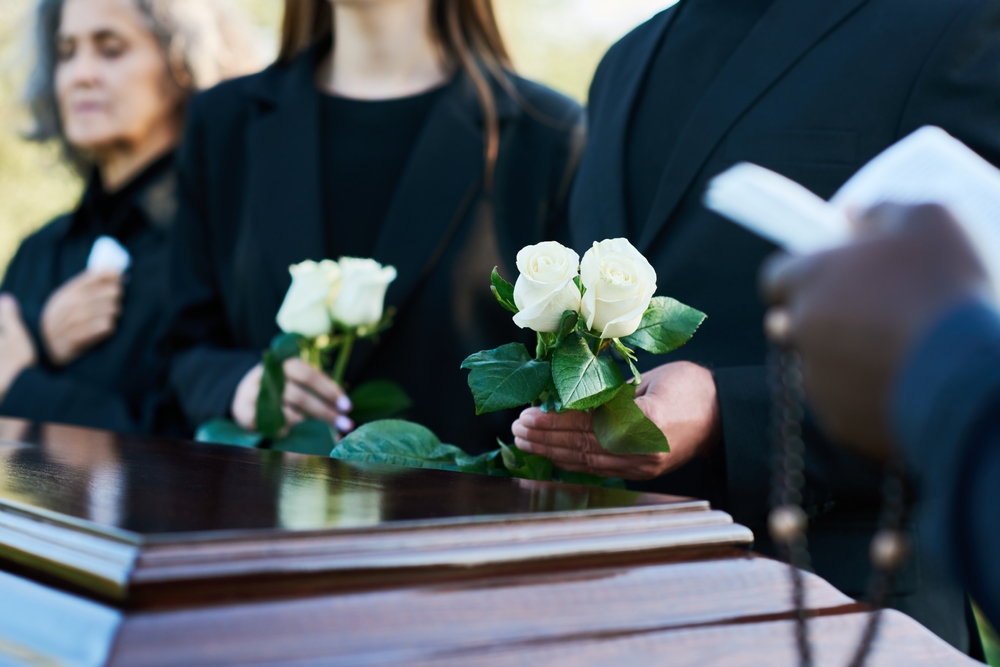 Hands, rose and a person at a funeral in a cemetery in grief while mourning loss at a memorial service. Death, flower and an adult in a suit at a graveyard in a crowd for an outdoor burial closeup
