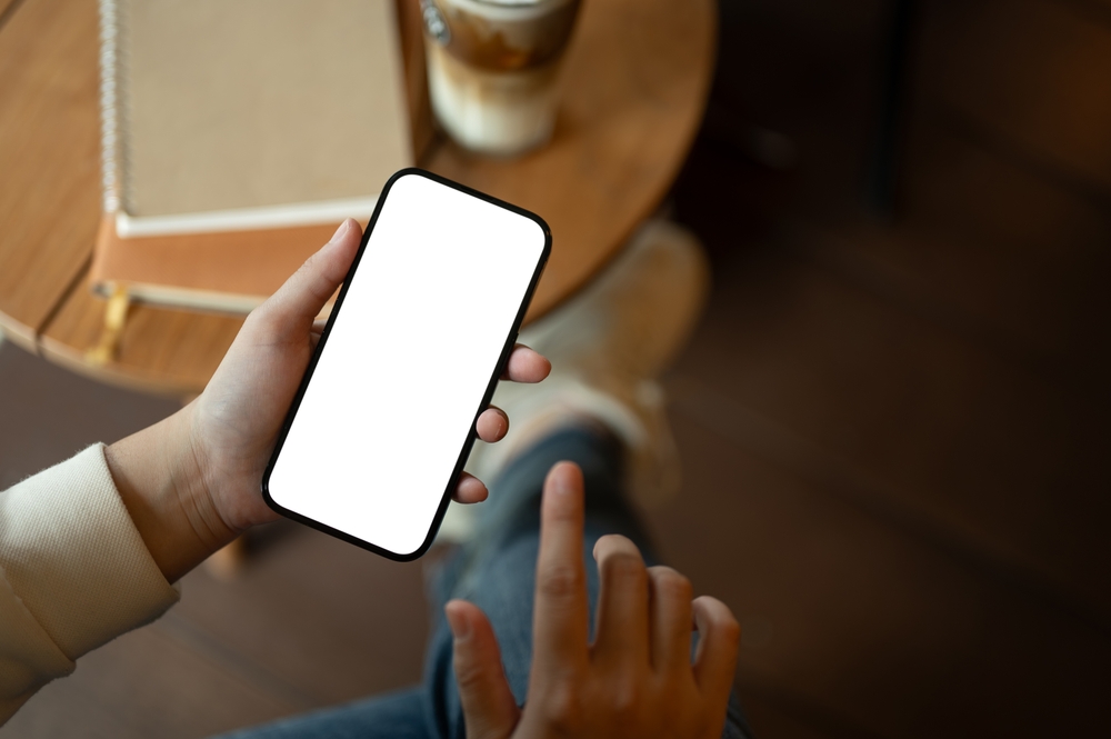 A woman is chilling in a cafe coffee shop and using her smartphone at a table while enjoying her coffee. A white-screen smartphone mockup for display your graphic ads. close-up image