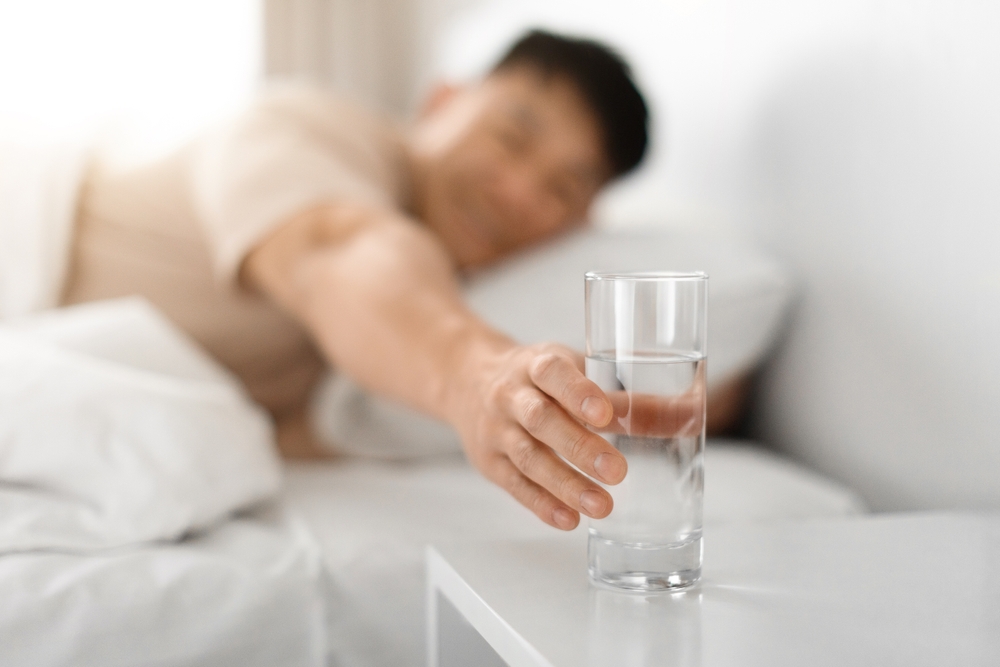 Healthy asian middle aged man lying in bed at home, reaching with hand glass, drinking water after waking up in the morning, copy space, blurred background